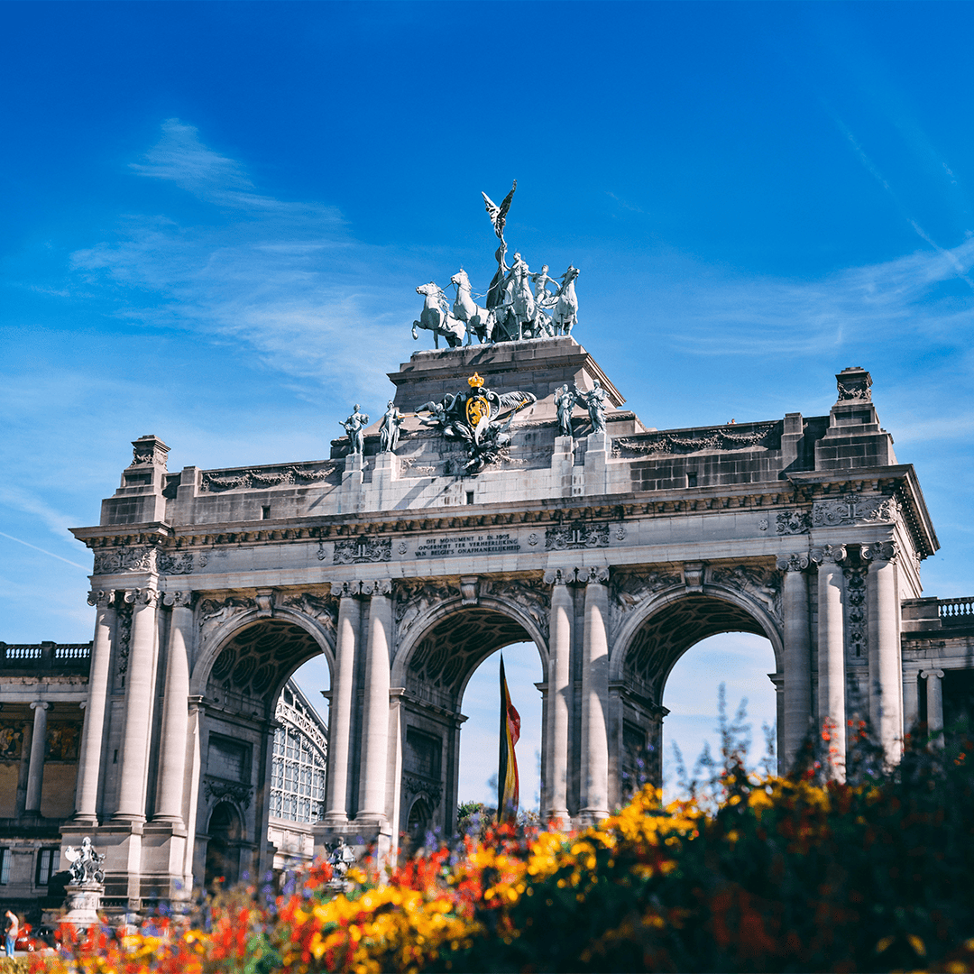 Triumphal arch with intricate sculptures and statues on top against a clear blue sky. Vibrant flowers in foreground.