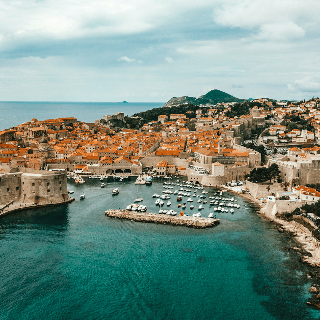 Aerial view of Dubrovnik's old town, featuring orange rooftops, historic stone walls, and a harbor with boats, set against a turquoise sea.