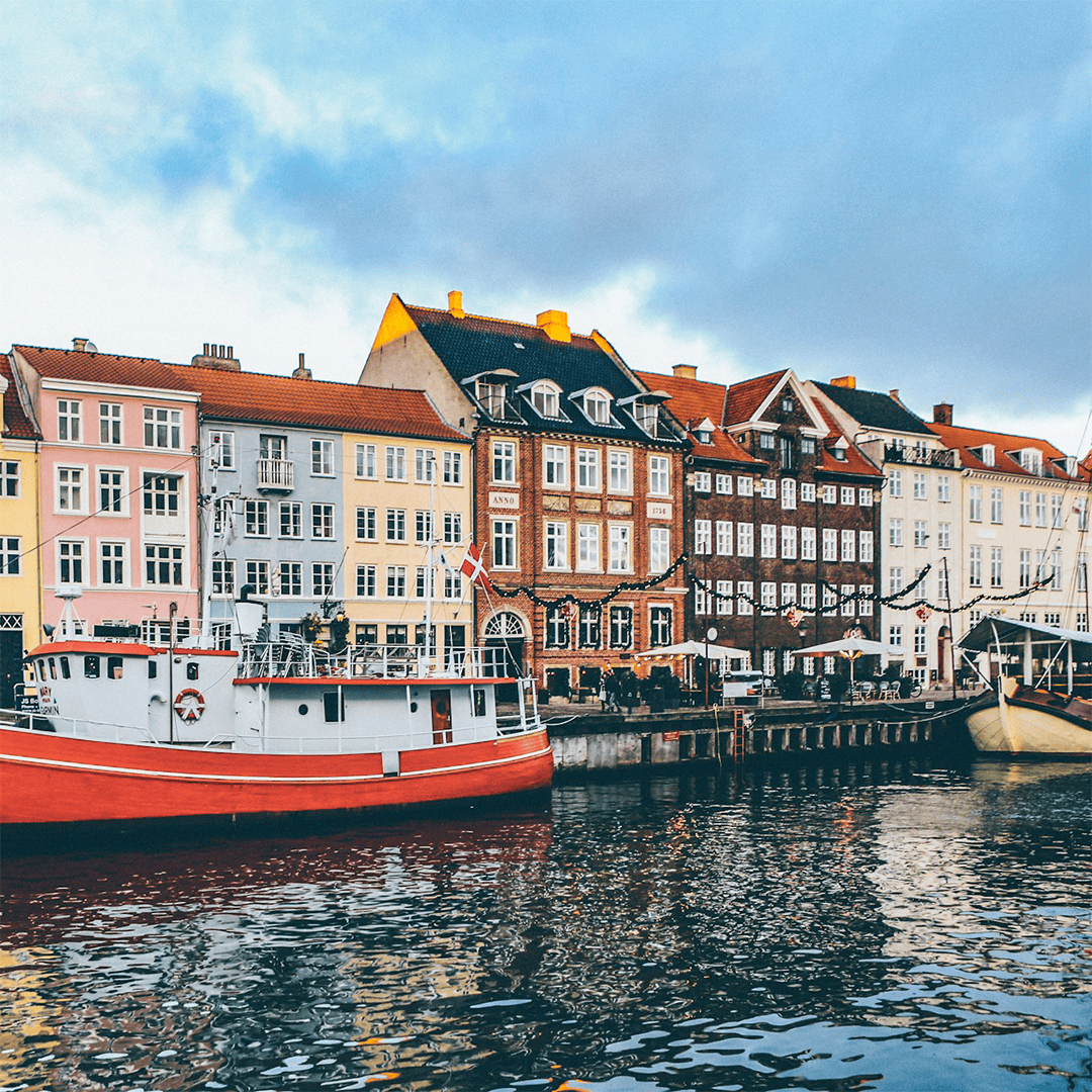 Colorful, historic buildings line a canal in Copenhagen, Denmark. A red and white boat is docked in calm water, under a cloudy, bright sky.