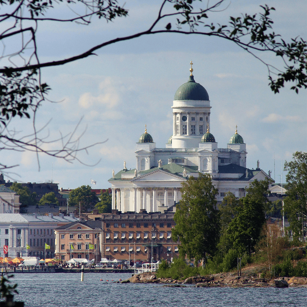 View of Helsinki Cathedral in Finland from across the water, framed by tree branches. The building's green domes stand out against a blue sky.