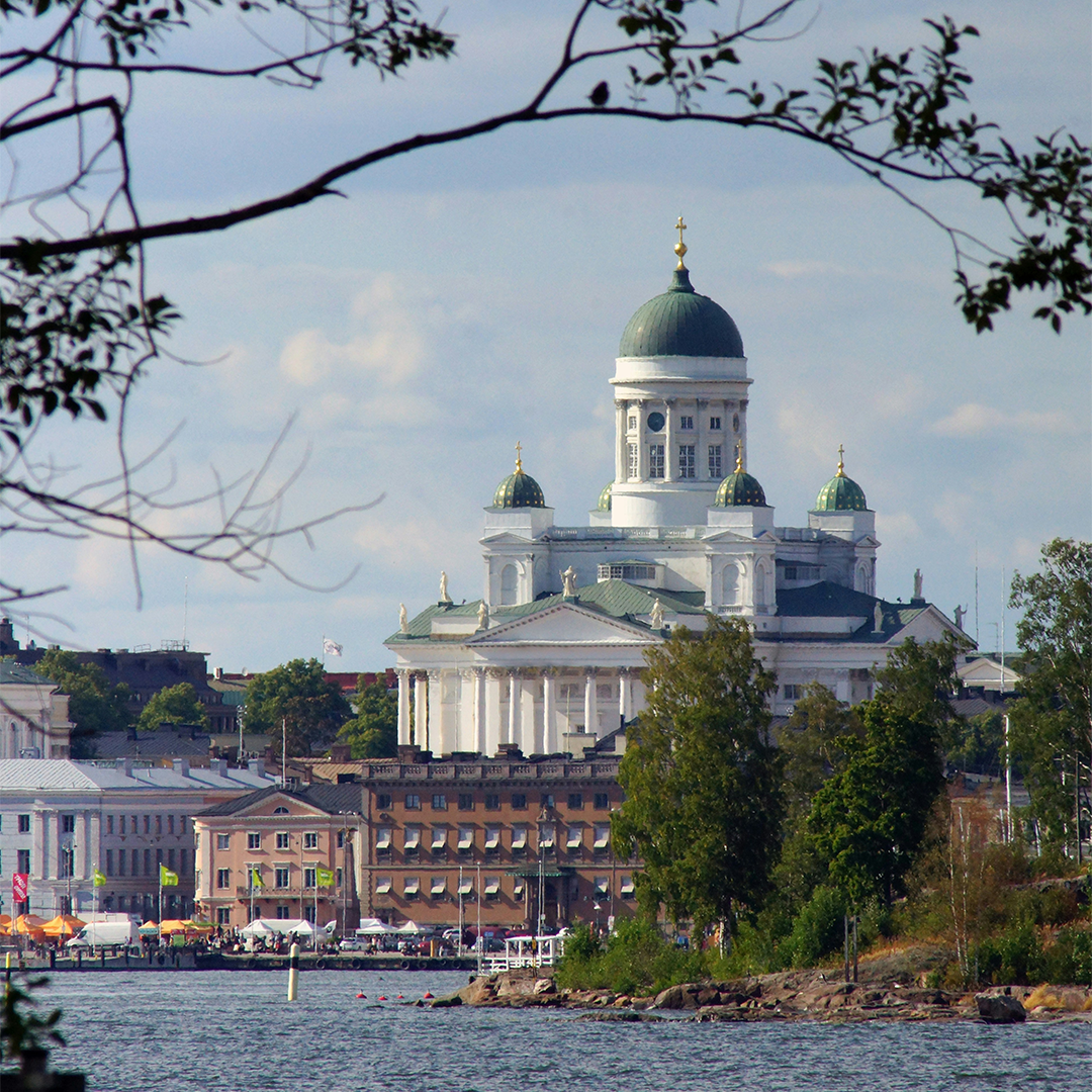 View of Helsinki Cathedral in Finland from across the water, framed by tree branches. The building's green domes stand out against a blue sky.