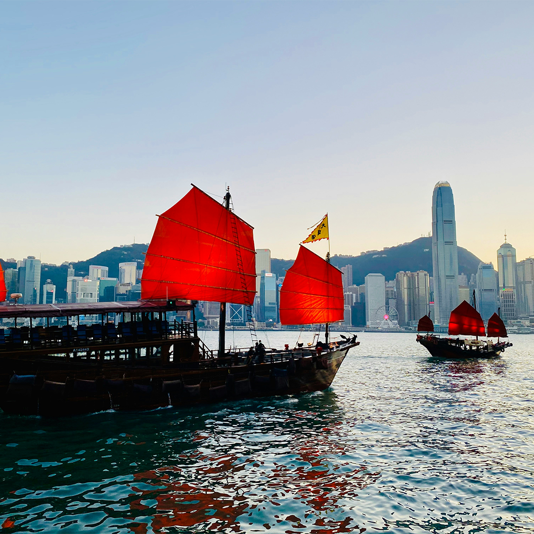 Traditional junk boats with red sails float on a calm harbor at sunset, against a modern city skyline and mountains.