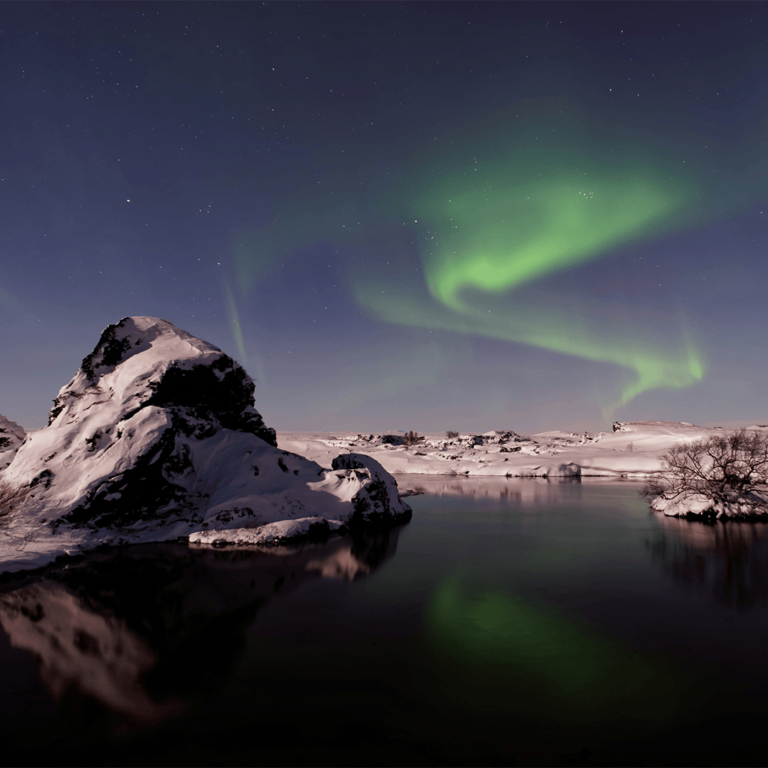 Snowy landscape with a rocky hill beside a calm lake under a night sky. The green Northern Lights illuminate the sky.