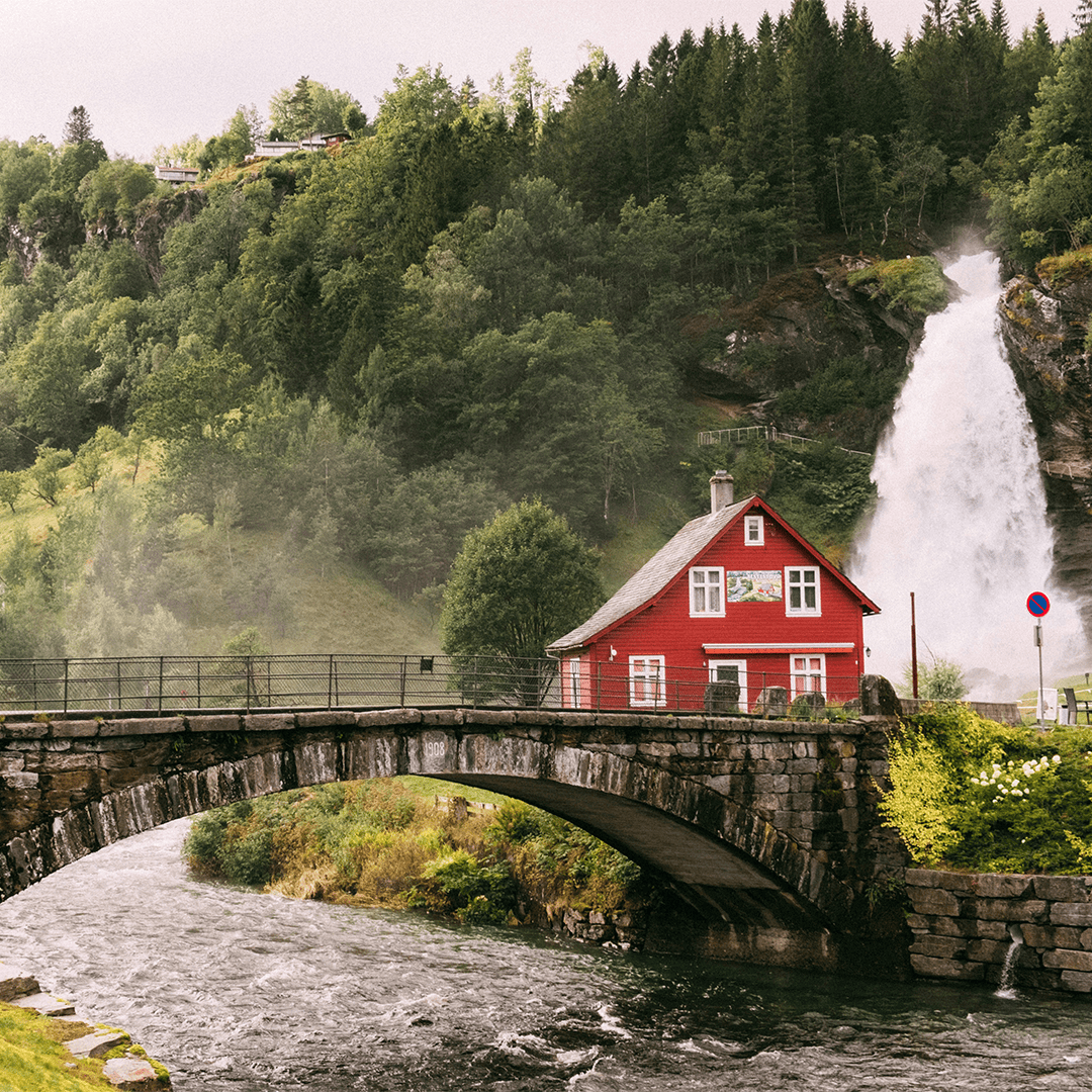 A charming red house sits beside a roaring waterfall and lush green forest. A stone bridge arches over a flowing river.