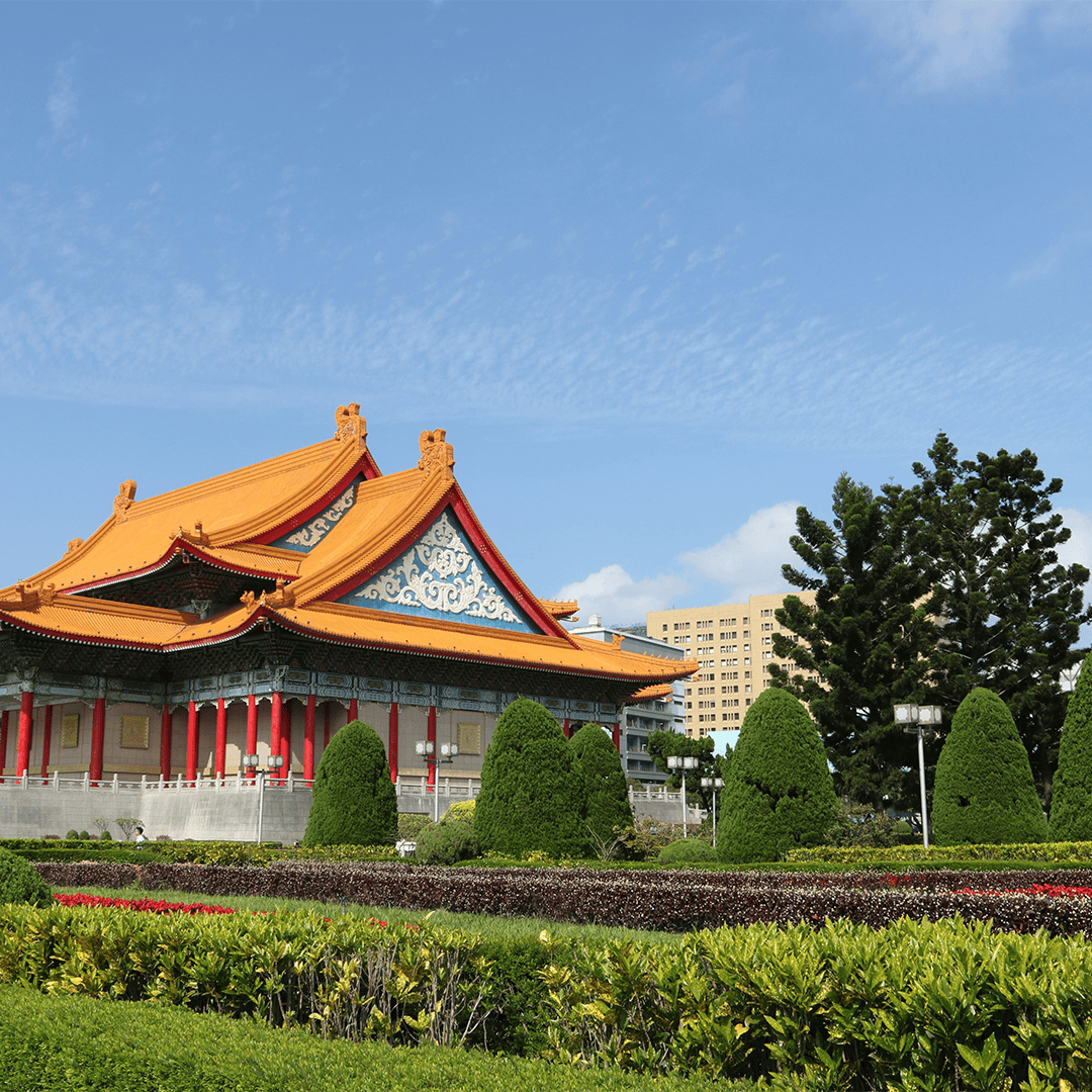 Traditional Asian architecture with vibrant orange roof and red columns, surrounded by manicured greenery and topiary against a clear blue sky.