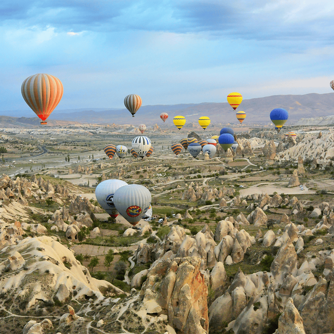 Hot air balloons in various colors float over the rocky landscape of Cappadocia under a blue sky.