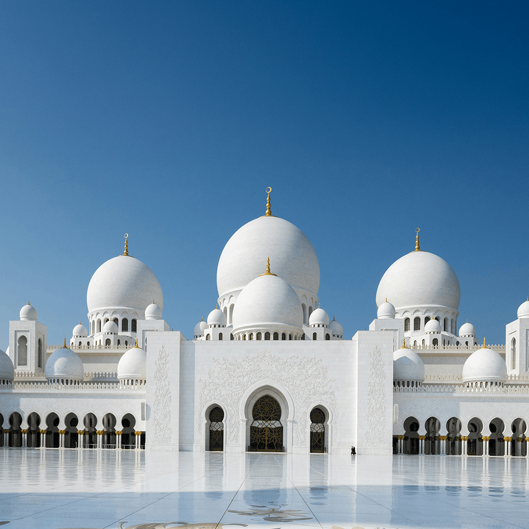 White mosque with multiple domes and arches, set against a clear blue sky.