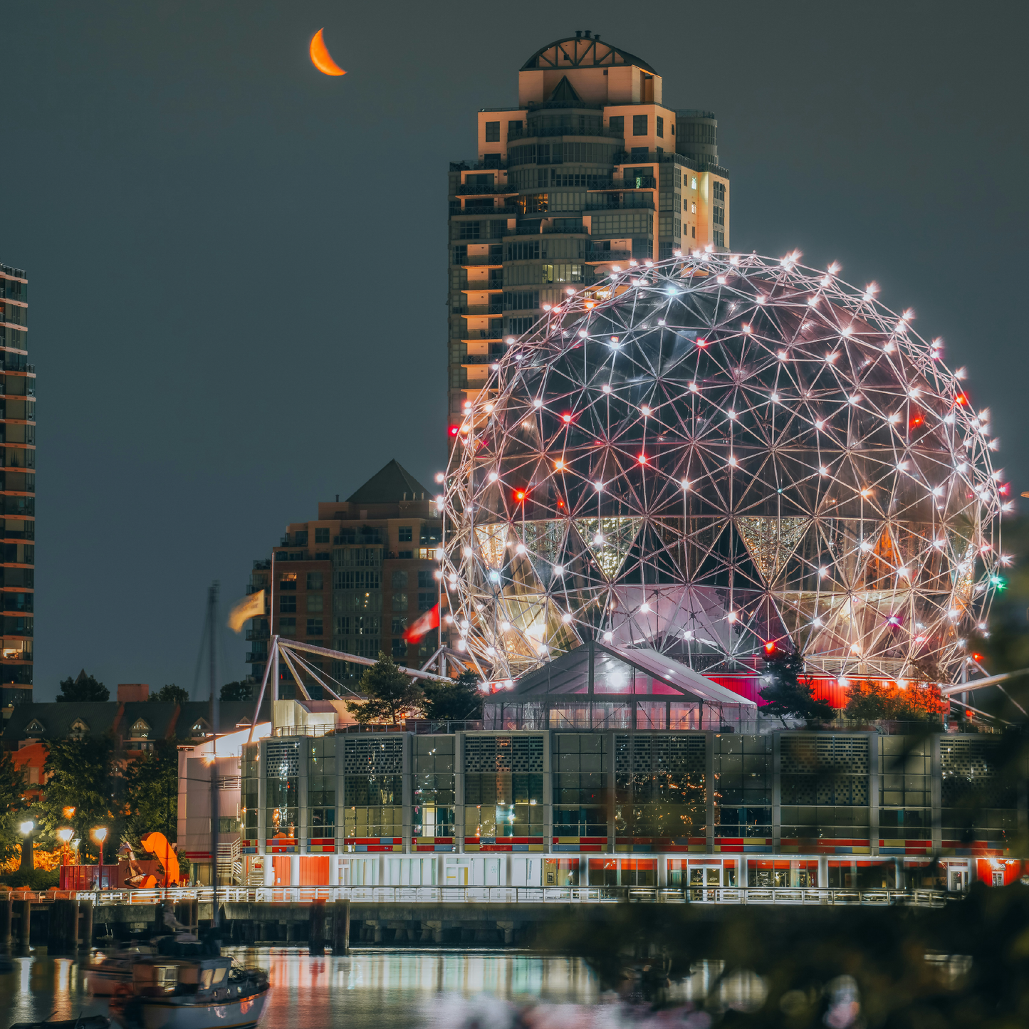 Science World, a geodesic dome illuminated with white and red lights at night, sits along the waterfront in Vancouver, Canada, with a crescent moon glowing orange in the dark sky above and city buildings visible in the background.