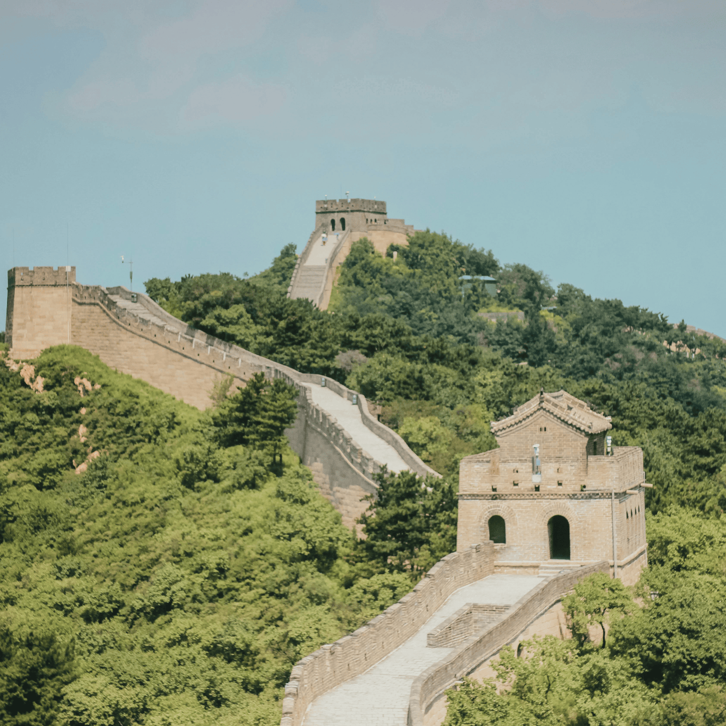 The Great Wall of China winds across forested mountain ridges, featuring stone watchtowers and crenellated ramparts under a pale blue sky.