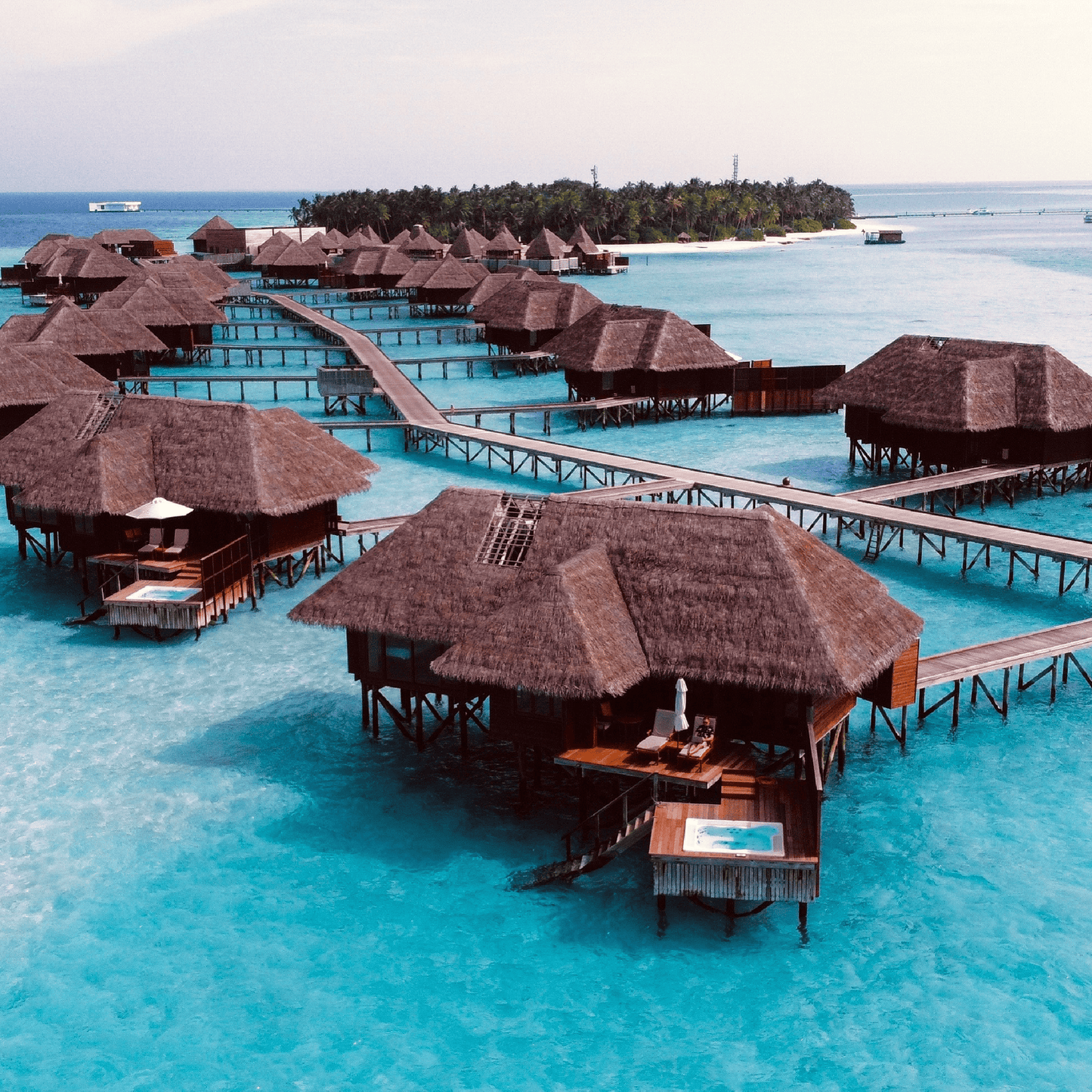 Aerial view of overwater bungalows with thatched roofs connected by wooden walkways over crystal-clear turquoise water, with a palm-fringed island in the distance.