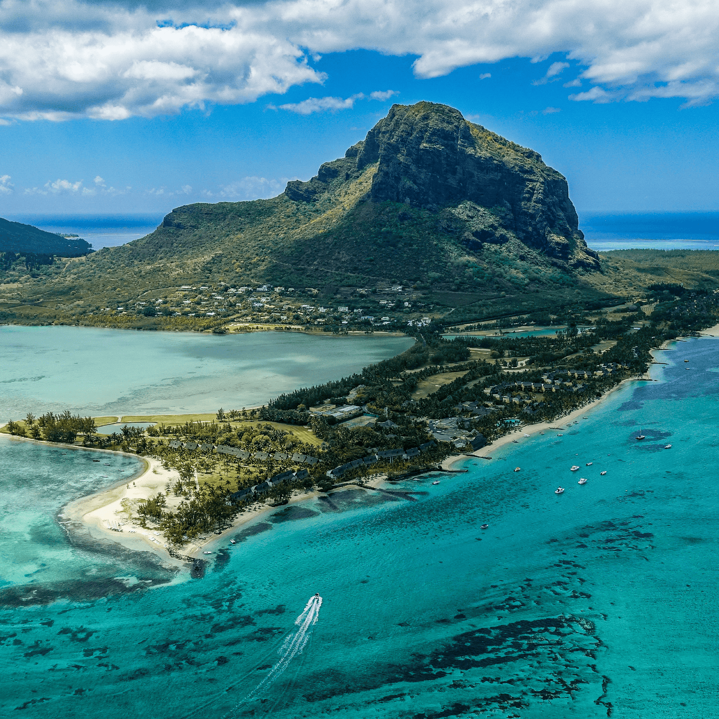 Aerial view of Le Morne Brabant mountain rising above turquoise lagoon waters, with sandy beaches, coastal vegetation, and small boats visible under a partly cloudy blue sky in Mauritius.