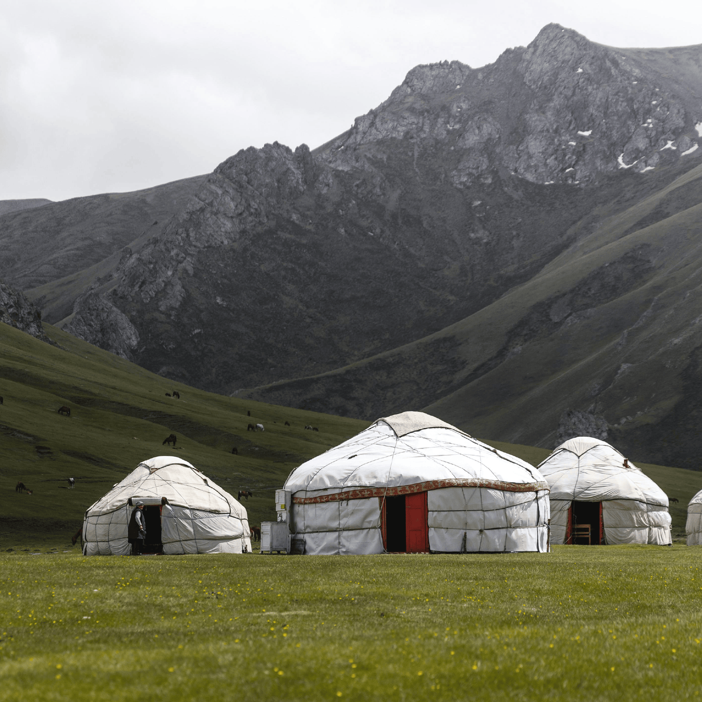 Three traditional white gers (yurts) with red doors stand on a green mountain meadow beneath a dramatic rocky peak with snow-capped ridges under an overcast sky.