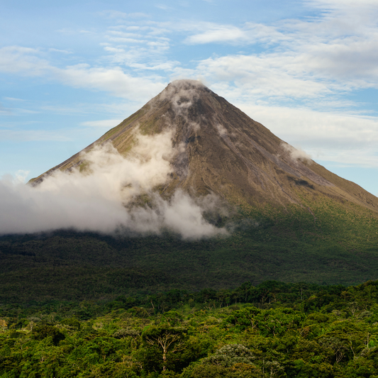 Arenal Volcano in Costa Rica covered in low clouds, rising above a dense tropical forest under a blue sky with scattered white clouds.
