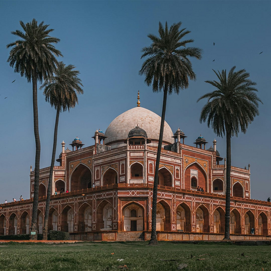 A grand red sandstone tomb with a white dome stands among tall palm trees, with birds flying against a bright blue sky.