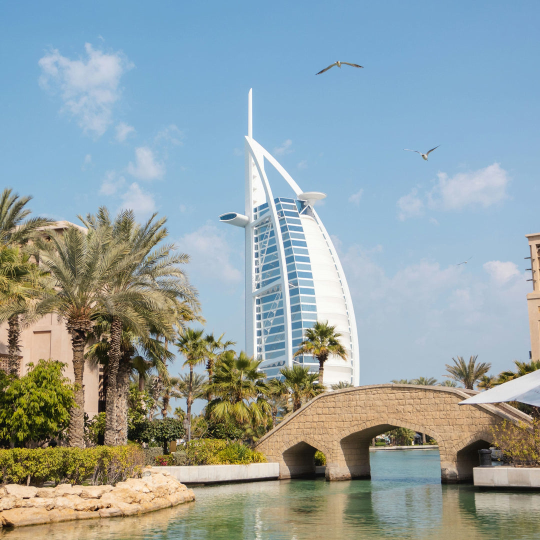 The Burj Al Arab rises above palm trees and landscaped gardens, with birds flying beneath a cloudy sky.