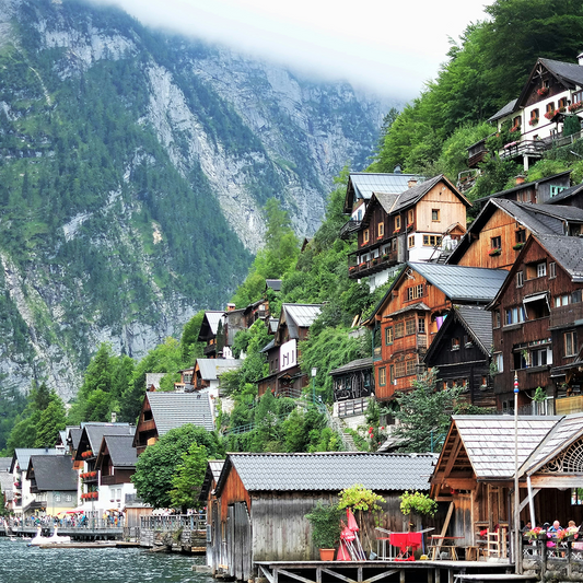 Alpine village scene with traditional wooden houses clinging to a lush, green hillside. A serene, mist-covered mountain looms in the background.