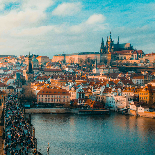 Scenic cityscape of a Czech city with a bustling crowd on a historic bridge over a river. Red-roofed buildings and a castle dominate the background under a vibrant blue sky.