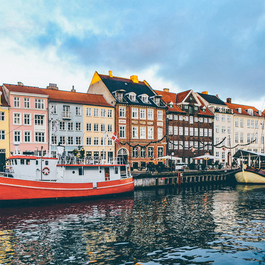 Colorful, historic buildings line a canal in Copenhagen, Denmark. A red and white boat is docked in calm water, under a cloudy, bright sky.