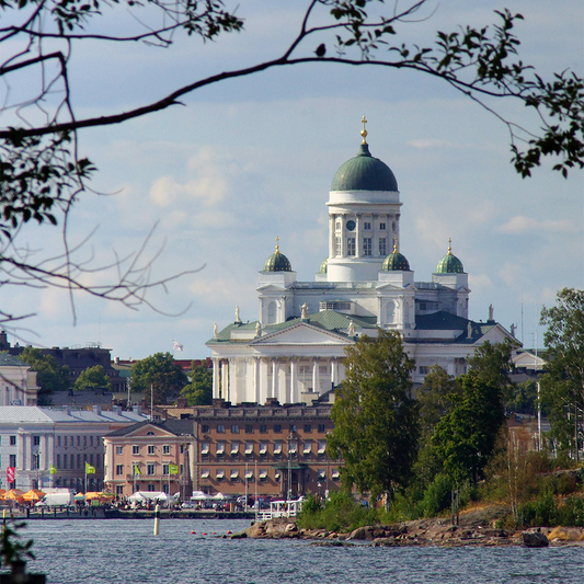 View of Helsinki Cathedral in Finland from across the water, framed by tree branches. The building's green domes stand out against a blue sky.