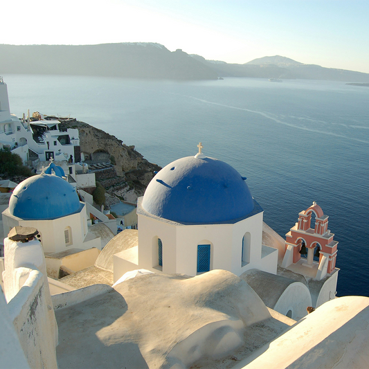 Scenic view of Greece, featuring iconic white buildings with blue domes overlooking the calm, expansive Aegean Sea under a clear sky.