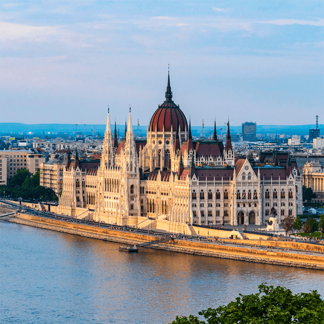The image shows the Budapest Parliament building at sunset, with ornate spires and a central dome, reflecting in the calm waters of the Danube River.