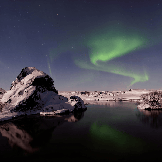 Snowy landscape with a rocky hill beside a calm lake under a night sky. The green Northern Lights illuminate the sky.