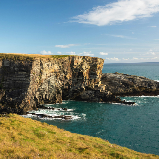 Coastal cliff view with rugged rock formations meeting the sea, under a blue sky with clouds. Lush grass covers the cliff top.