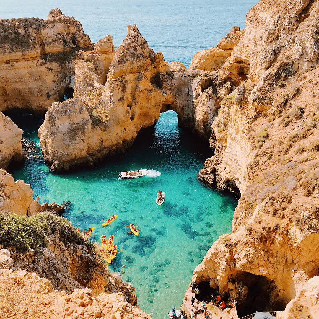Aerial view of a vibrant coastal scene. Turquoise waters surround rugged orange cliffs. People are kayaking and a boat sails.
