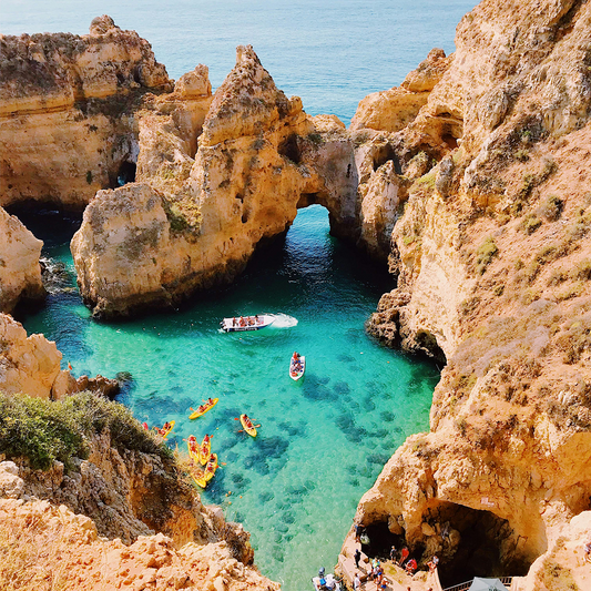 Aerial view of a vibrant coastal scene. Turquoise waters surround rugged orange cliffs. People are kayaking and a boat sails.