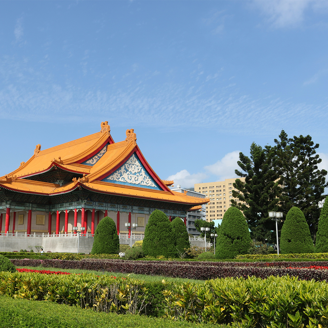 Traditional Asian architecture with vibrant orange roof and red columns, surrounded by manicured greenery and topiary against a clear blue sky.