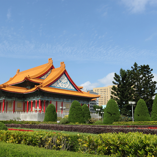 Traditional Asian architecture with vibrant orange roof and red columns, surrounded by manicured greenery and topiary against a clear blue sky.
