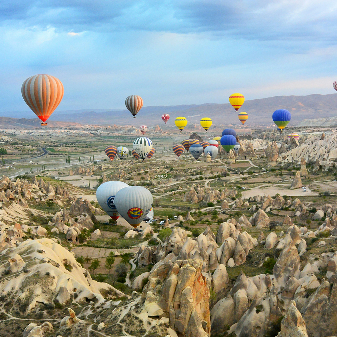 Hot air balloons in various colors float over the rocky landscape of Cappadocia under a blue sky.