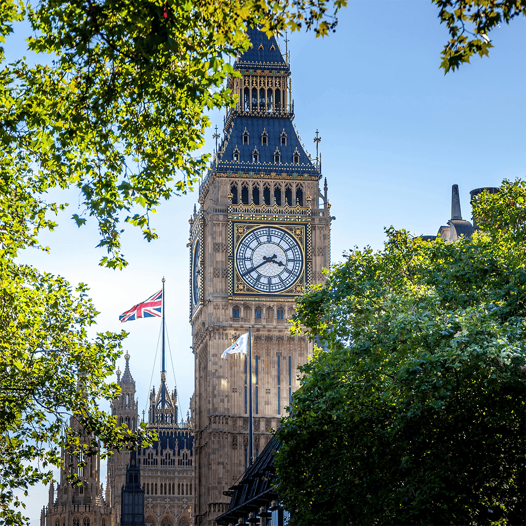 The image shows Big Ben against a clear blue sky, surrounded by green trees. The Union Jack flag flies prominently, conveying a bright, iconic London scene.