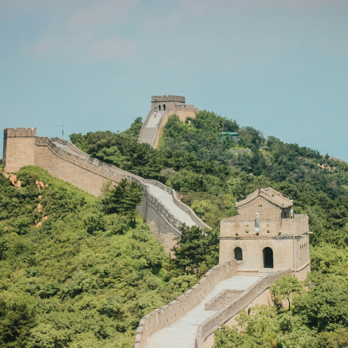 The Great Wall of China winds across forested mountain ridges, featuring stone watchtowers and crenellated ramparts under a pale blue sky.