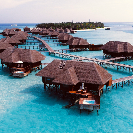 Aerial view of overwater bungalows with thatched roofs connected by wooden walkways over crystal-clear turquoise water, with a palm-fringed island in the distance.