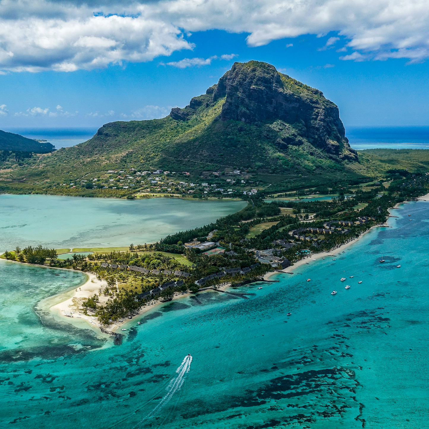 Aerial view of Le Morne Brabant mountain rising above turquoise lagoon waters, with sandy beaches, coastal vegetation, and small boats visible under a partly cloudy blue sky in Mauritius.