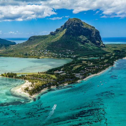 Aerial view of Le Morne Brabant mountain rising above turquoise lagoon waters, with sandy beaches, coastal vegetation, and small boats visible under a partly cloudy blue sky in Mauritius.