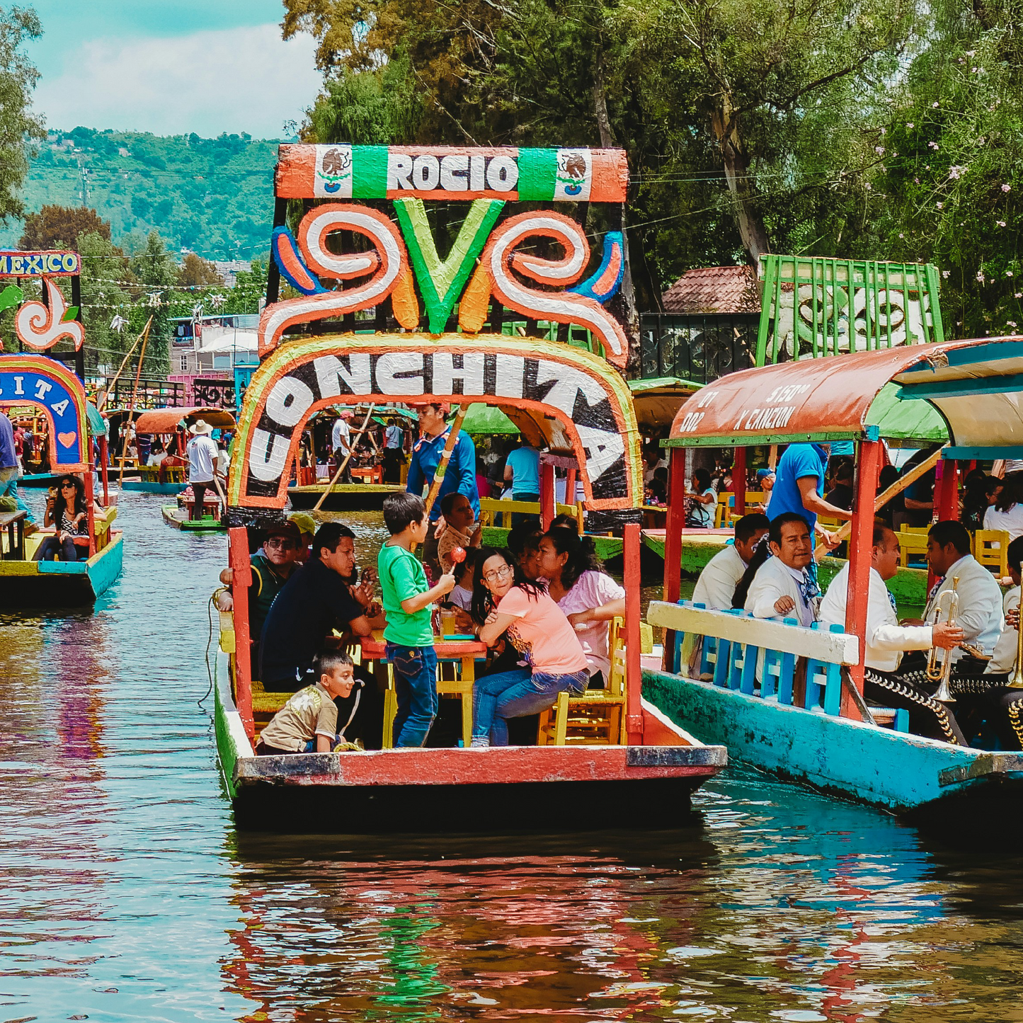 Colourful trajineras (decorated boats) filled with passengers navigate a canal in Xochimilco, Mexico, with vibrant hand-painted signage, mariachi musicians, and lush trees lining the waterways.