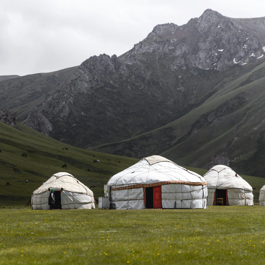 Three traditional white gers (yurts) with red doors stand on a green mountain meadow beneath a dramatic rocky peak with snow-capped ridges under an overcast sky.