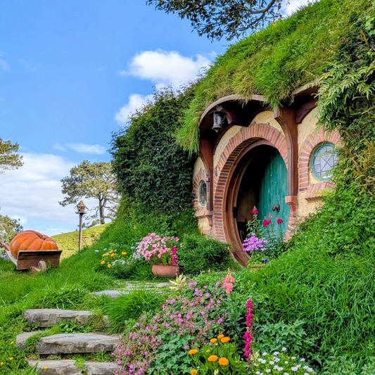 A vibrant green hobbit house with a round teal door and brick archway, built into a grassy hillside at Hobbiton in New Zealand, surrounded by colourful flower gardens, stone steps, and a wooden wheelbarrow holding a giant pumpkin under a bright blue sky.