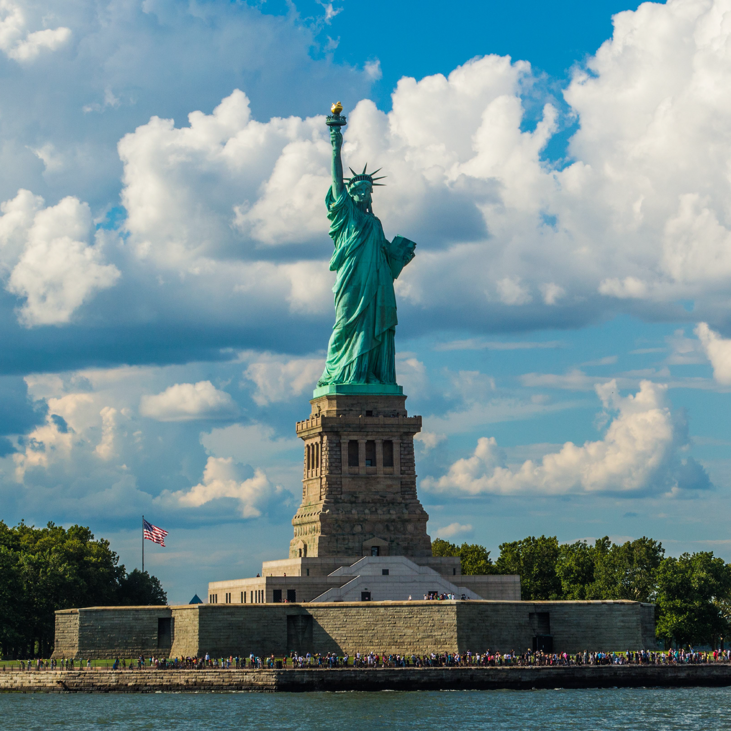 The Statue of Liberty stands on its stone pedestal against a bright blue sky with scattered white clouds, surrounded by green trees and crowds of visitors along the waterfront in New York Harbor.