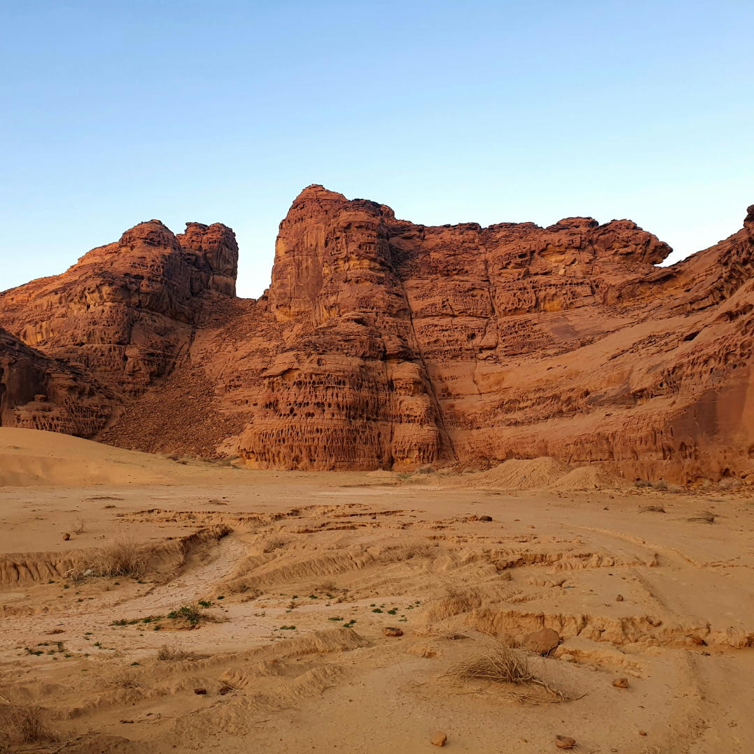A scenic desert landscape in Saudi Arabia with layered orange-brown rock formations rising above sandy dunes under a clear pale blue sky.