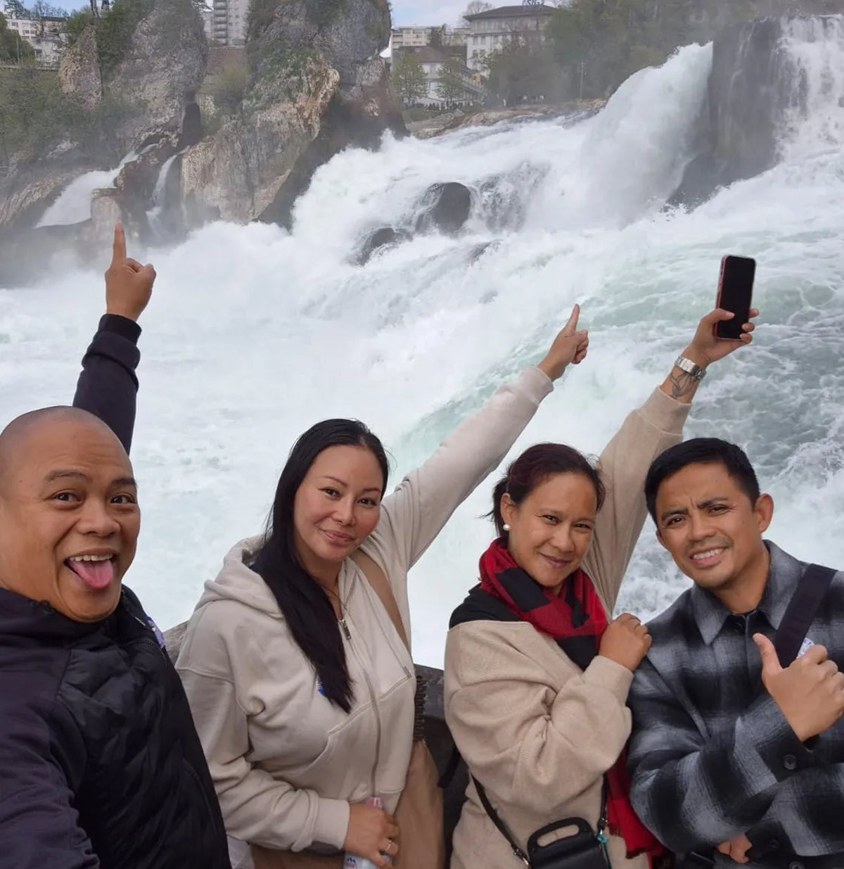 Four people taking a selfie in front of a large waterfall, pointing upward and smiling. The group is dressed in casual outdoor clothing, with the rushing water and rocky cliffs filling the background.